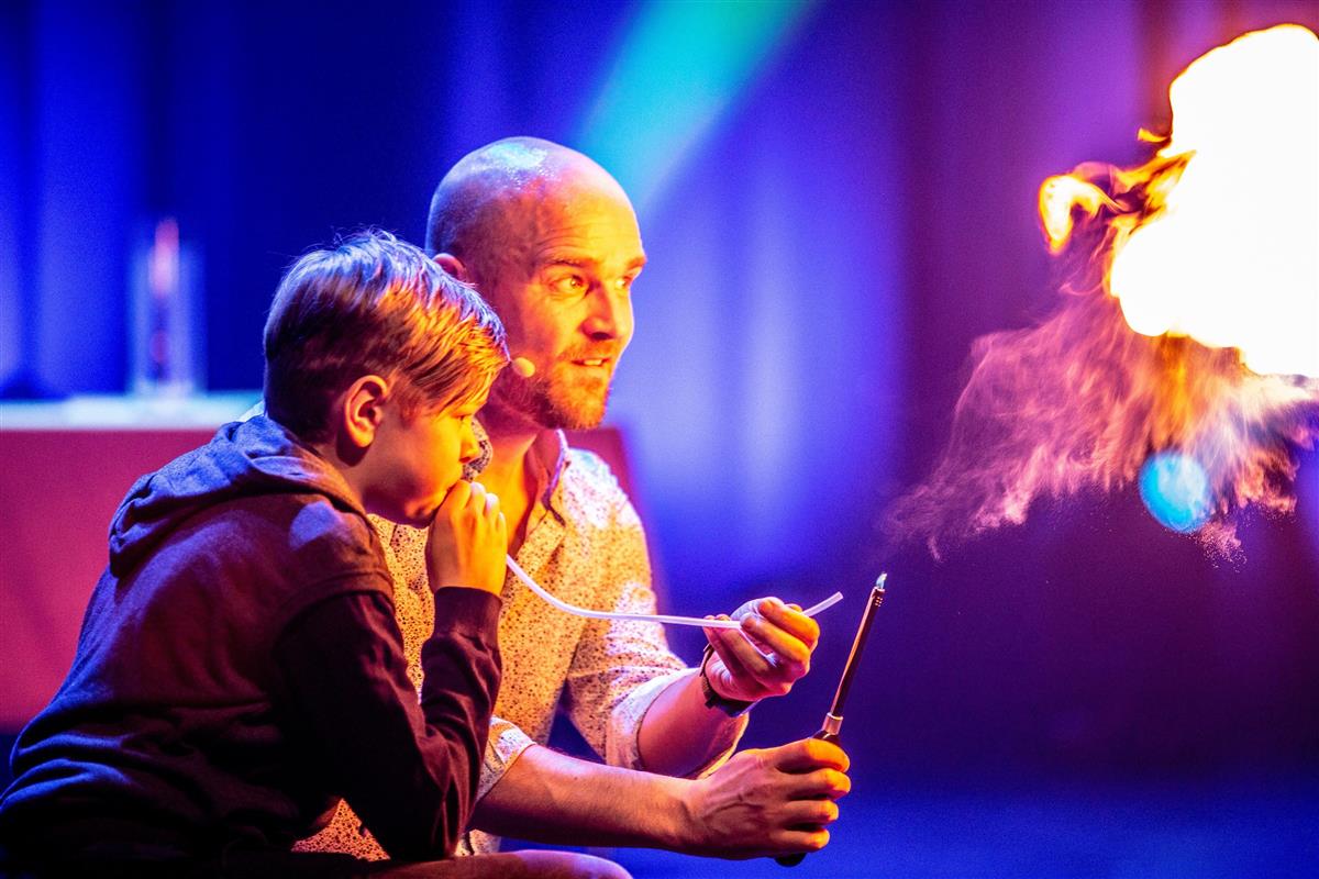 Håvard og eit barn sit på huk på ein scene og blås flammar